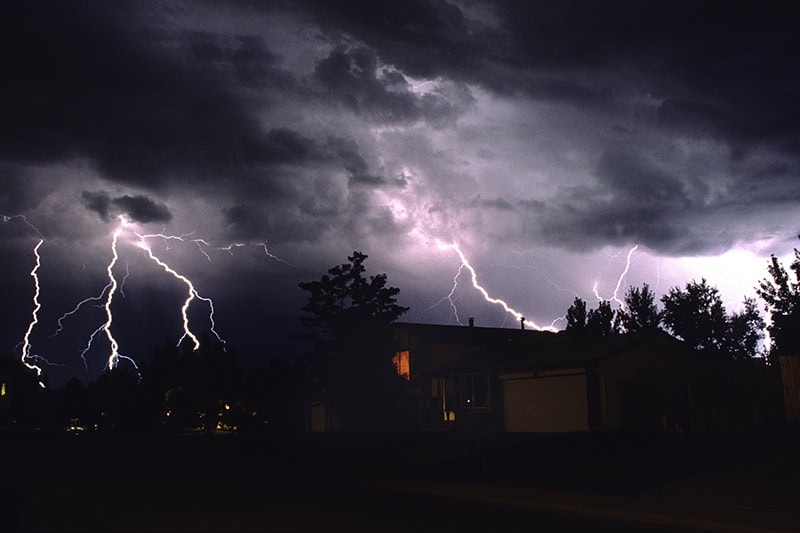 As rain falls, lightning bolts strike over southwest suburban Denver, Colorado. An window is lit dimly in the middle of the silhouetted trees.