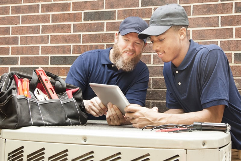 Multi-ethnic team of men repairing a home's air conditioner unit outdoors. They use a digital tablet to access the repairs.  They both wear blue uniforms.